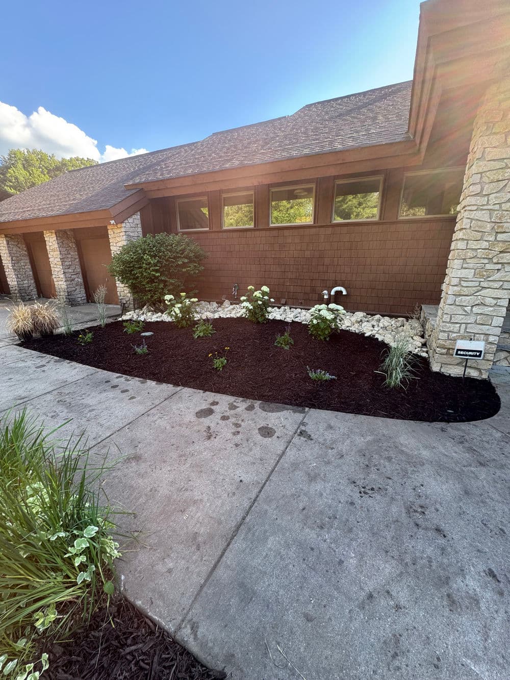 Modern home landscape with black mulch, flowering plants, and stone features under clear blue sky.