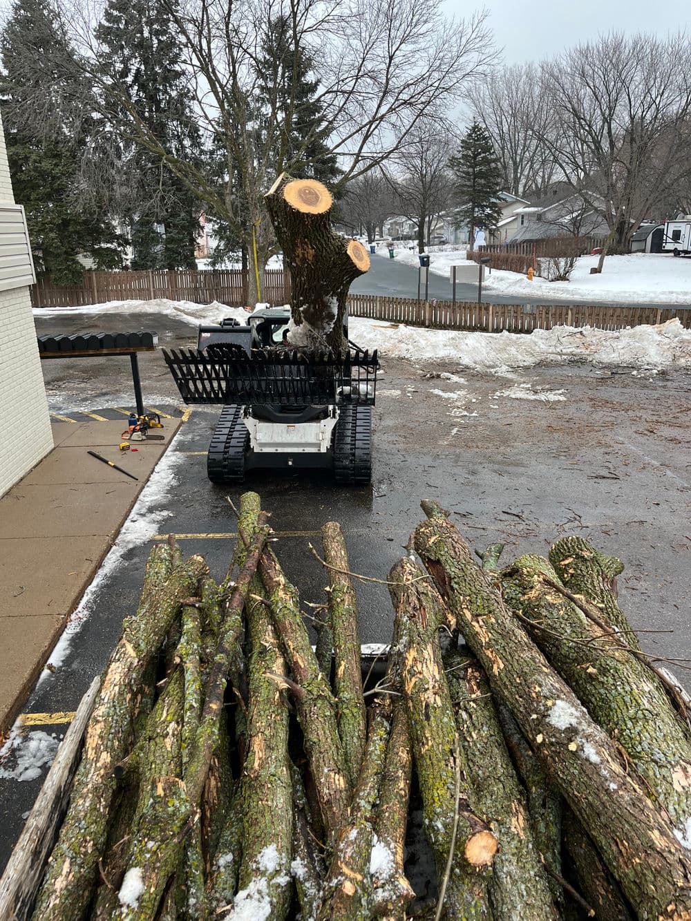Tree service equipment cutting logs in a snowy parking lot with stacked branches nearby.