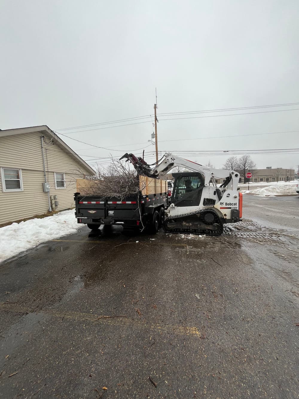 Bobcat loader unloading branches from a trailer in a snowy urban setting.