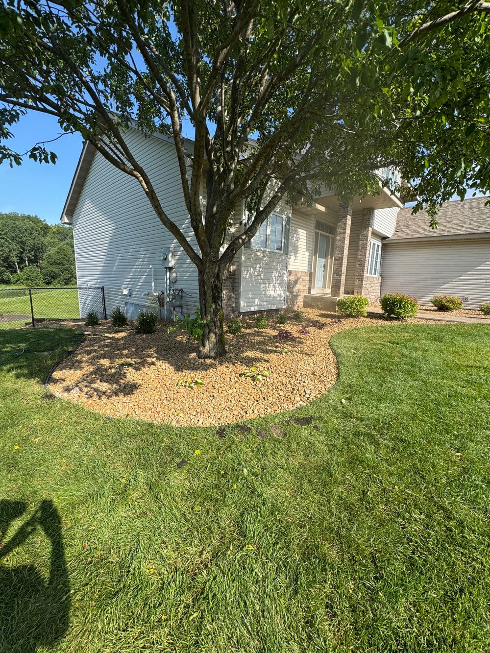 Landscaped home exterior with tree, stone border, and green lawn on a sunny day.